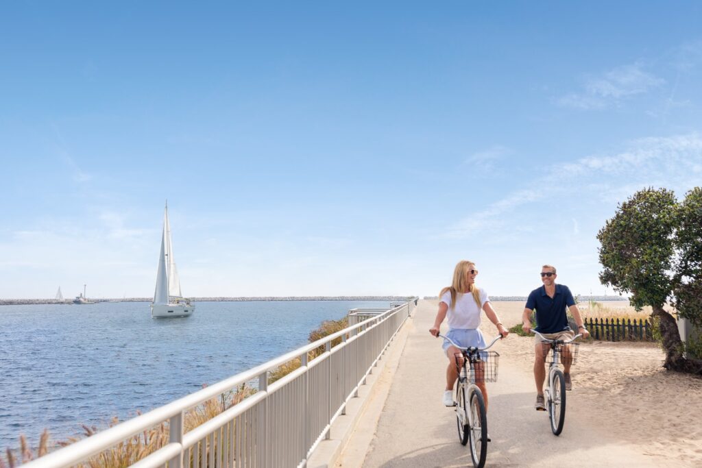 Two people riding bikes on paved path next to ocean and sailboat