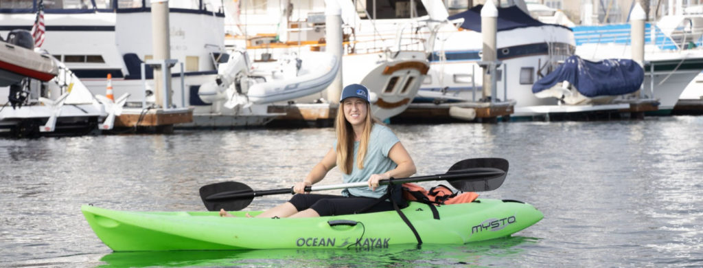 woman paddling in a green kayak in the sea with boats docked nearby